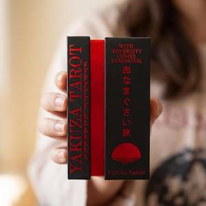 May include: A black and red tarot card deck box featuring the text YAKUZA TAROT on the side and WITH ADVERSITY COMES STRENGTH with Japanese characters and a red sun graphic on the front. The box has a vibrant red spine visible between two black panels.