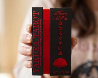 May include: A black and red tarot card deck box featuring the text YAKUZA TAROT on the side and WITH ADVERSITY COMES STRENGTH with Japanese characters and a red sun graphic on the front. The box has a vibrant red spine visible between two black panels.