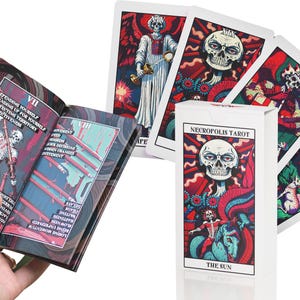 May include: A set of tarot cards with a dark, gothic aesthetic. The cards feature illustrations of skeletons, skulls, and other macabre imagery. The box for the cards is white with black text that reads "Necropolis Tarot" and "The Sun".