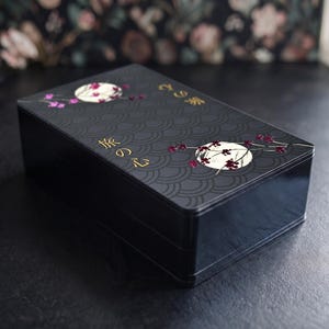 May include: A rectangular black box with a matte finish featuring a repeating dark gray wave pattern. The lid displays two white circular motifs with dark pink floral branches and gold Japanese text characters reading 旅の心.