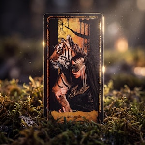 May include: A rectangular tarot card labeled Strength with the Roman numeral VIII, featuring an illustration of a person wearing a gold mask standing beside a tiger. The card has a dark border and a textured, weathered-looking bottom section.