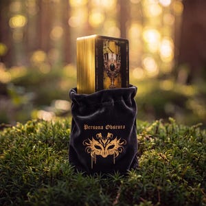 May include: A deck of tarot cards with metallic gold edges partially inside a black velvet drawstring pouch. The pouch features the text Persona Obscura and a gold graphic of a masquerade mask printed on the front surface of the fabric.
