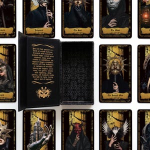 May include: A black rectangular storage box with a hinged lid, featuring gold text and a mask emblem on the interior. The box is surrounded by multiple tarot cards displaying dark, gothic-style illustrations of figures in masks, robes, and ornate headpieces.