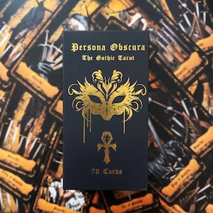 May include: A black rectangular tarot deck box with gold foil lettering that reads Persona Obscura The Gothic Tarot. The box features a gold foil mask illustration and an ankh symbol, with the text 78 Cards printed at the bottom.