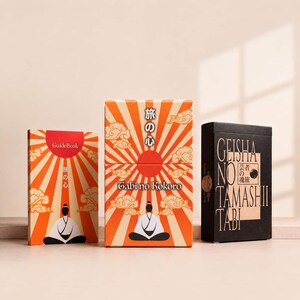 May include: Two rectangular boxes and a guidebook featuring orange and cream sunburst patterns with stylized clouds and a figure in a seated pose. The central box displays the text Tabi no Kokoro, while the black box is labeled Geisha no Tamashii Tabi.