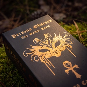 May include: A black rectangular tarot deck box with gold foil lettering and graphics. The text reads Persona Obscura The Gothic Tarot. A gold ornate masquerade mask illustration is centered above a gold ankh symbol on the front of the box.