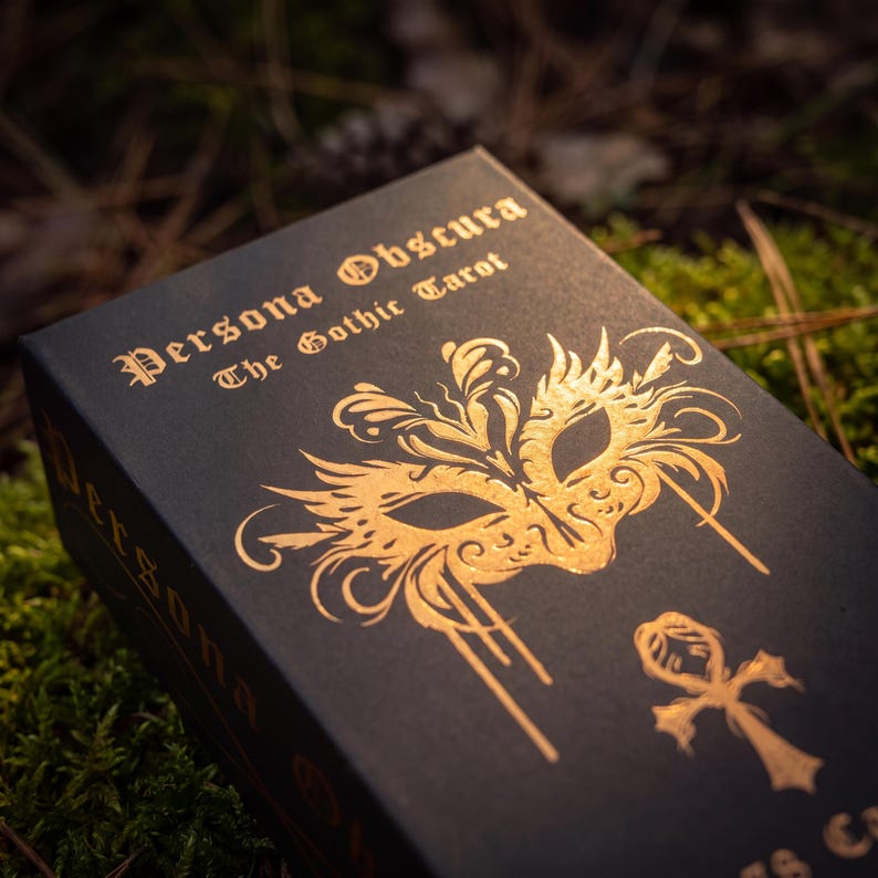 May include: A black rectangular tarot deck box with gold foil lettering and graphics. The text reads Persona Obscura The Gothic Tarot. A gold ornate masquerade mask illustration is centered above a gold ankh symbol on the front of the box.