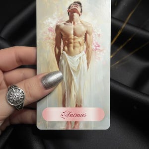 Puede incluir: Carta del tarot con una figura masculina musculosa envuelta en tela blanca, con la palabra "Animus" en la parte inferior. El fondo tiene suaves tonos rosas y blancos. Se ve un anillo plateado con un diseño de sol.