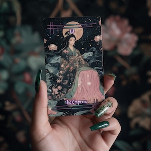 May include: A rectangular tarot card titled The Empress featuring an illustration of a figure in a dark floral robe with a pink skirt, set against a night sky with a large moon, stars, and floating flowers, framed by thin horizontal lines.