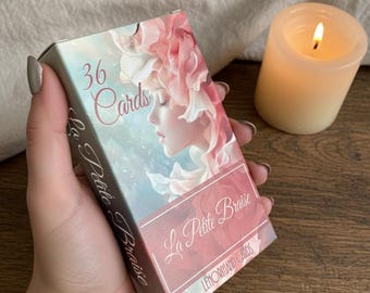Puede incluir: Una baraja de 36 cartas en una caja con el texto "La Petite Braise" y "36 Cards". La caja presenta una ilustración de acuarela de una mujer con acentos florales rosas. Una vela encendida está en el fondo.