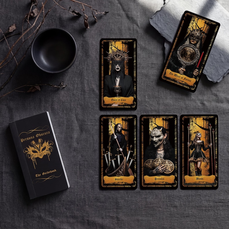 May include: A tarot deck set featuring a black guidebook titled Persona Obscura with a gold mask graphic and five rectangular tarot cards. The cards display dark, detailed illustrations of figures in masks and robes with gold and black color schemes.