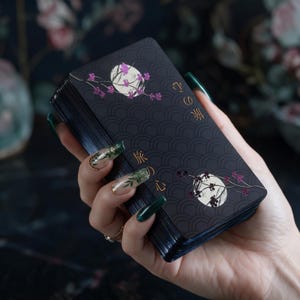 May include: A deck of rectangular cards with a matte black background featuring a repeating wave pattern, two white circular moons, pink floral branches, and gold Japanese text characters that read 旅の心, arranged in a vertical orientation.