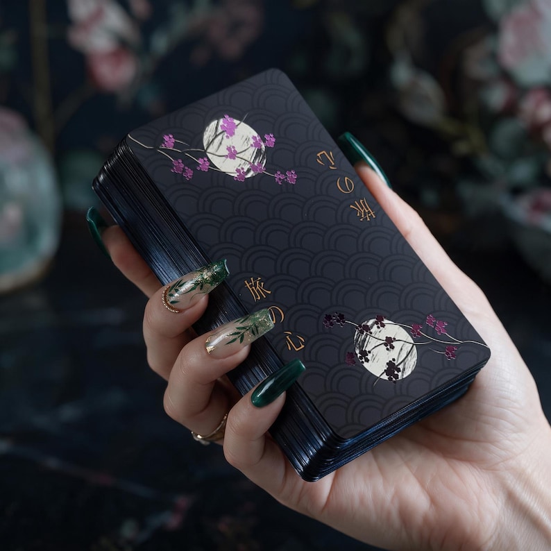 May include: A deck of rectangular cards with a matte black background featuring a repeating wave pattern, two white circular moons, pink floral branches, and gold Japanese text characters that read 旅の心, arranged in a vertical orientation.