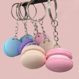 May include: Five colorful macaron keychains with silver key rings and chains. The macarons are in shades of blue, purple, beige, and pink.