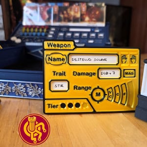 May include: A yellow game component with black trim, labeled "Weapon" and "DESTELLO SOLAR." It includes text fields for "Trait," "Damage," and "Range," along with a circular symbol and a logo. The component is on a wooden surface.