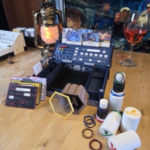 May include: A tabletop scene featuring a blue game organizer, game cards, and various game accessories. A vintage-style lantern with a lit bulb sits nearby. Several white cylindrical containers with colored accents are also present.