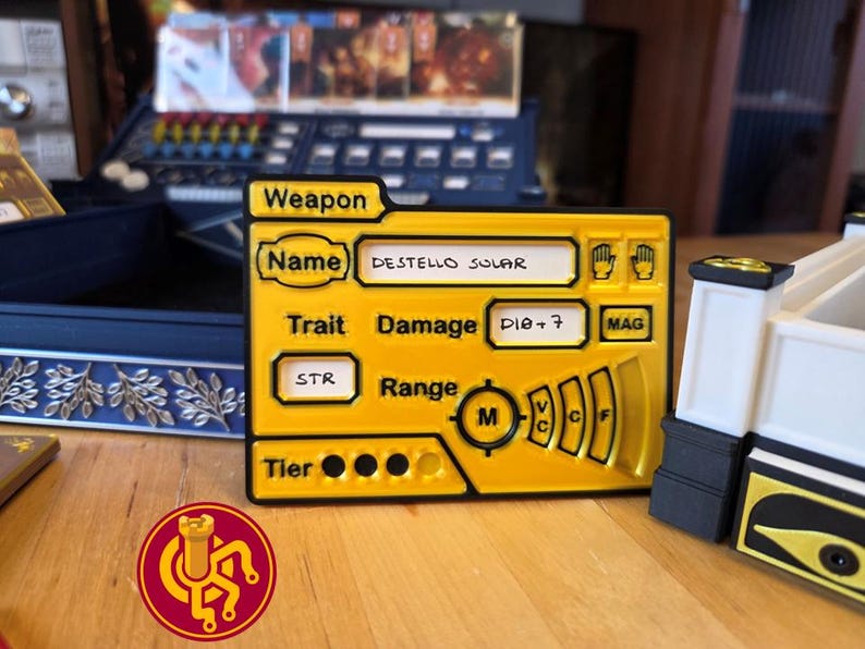 May include: A yellow game component with black trim, labeled "Weapon" and "DESTELLO SOLAR." It includes text fields for "Trait," "Damage," and "Range," along with a circular symbol and a logo. The component is on a wooden surface.