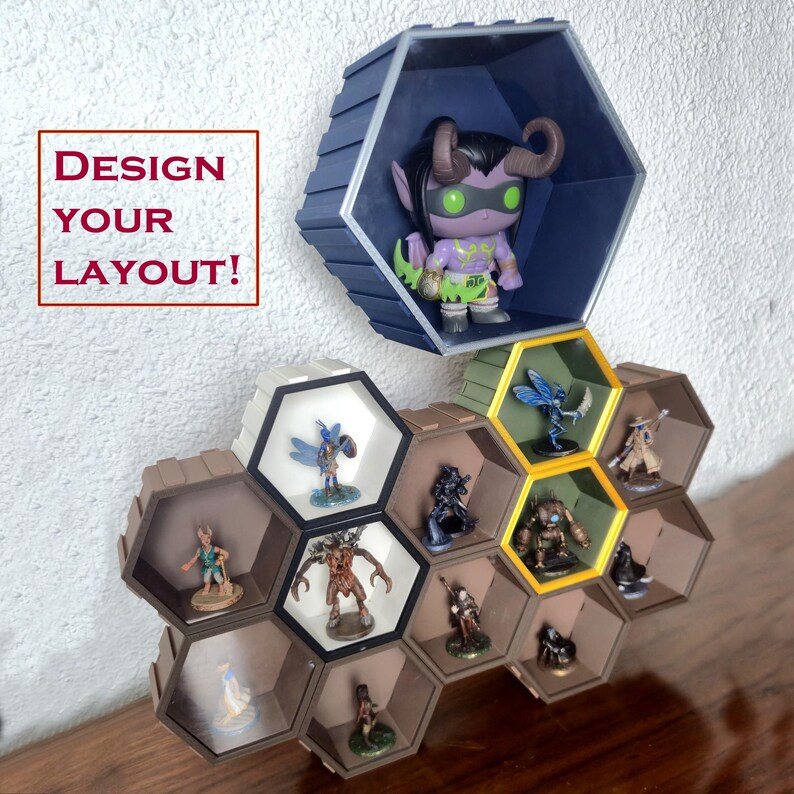 May include: Hexagonal display shelves in brown, blue, and yellow, showcasing various figurines. The shelves are mounted on a white wall, with the text "DESIGN YOUR LAYOUT!" in a red box. The shelves are designed to hold collectibles.