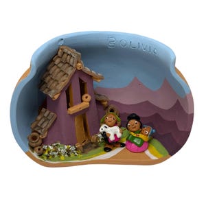 Puede incluir: Diorama de cerámica que representa una casa morada con techo marrón y tres figuras con vestimenta tradicional. El diorama está ambientado sobre un fondo de montañas y cielo azul, con la inscripción "BOLIVIA".