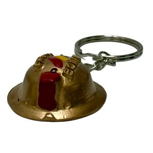 Könnte beinhalten: Ein goldfarbener Helm-Schlüsselanhänger mit einem silbernen Schlüsselring. Der Helm hat rote und gelbe Akzente, und das Wort "BOB" ist sichtbar. Ein kleines, dekoratives Objekt.
