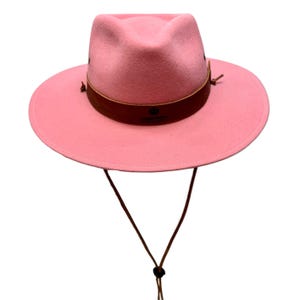 Op de afbeelding: Een roze fedora-hoed met een brede rand en een bruine leren band. De hoed heeft een kinband en is gemaakt van een zacht, viltachtig materiaal. De hoed is een stijlvol accessoire voor bescherming tegen de zon.