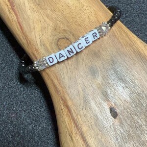 May include: A black beaded bracelet with white letter beads spelling "DANCER". The bracelet also features clear, faceted beads. The bracelet is displayed on a wooden surface.