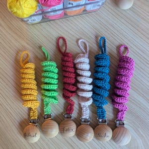 Peut inclure: Des attaches-tétines pour bébés faites à la main. Chaque attache comprend une perle en bois avec un nom, reliée à une spirale crochetée jaune, verte, rouge, blanche, bleue et violette. Un récipient transparent contient des pelotes de laine.