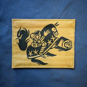 Op de afbeelding: Een rechthoekige patch met een zwarte illustratie op een beige achtergrond. De illustratie toont een figuur die op een gestileerd dier rijdt. De patch is geplaatst op een donkerblauwe stof. Het ontwerp heeft een vintage stijl.