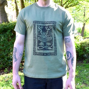 Könnte beinhalten: Ein grünes T-Shirt mit einem schwarz-weißen Grafikdruck eines Wesens mit Geweih. Das Wesen ist von einem Kettengliederrand umgeben.