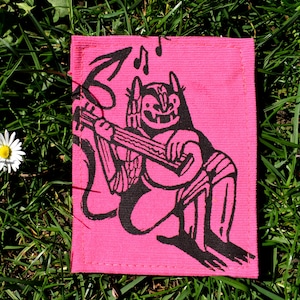 May include: A pink rectangular patch with a black illustration of a devil playing a stringed instrument, and musical notes. The patch is on green grass, with a daisy in the corner.