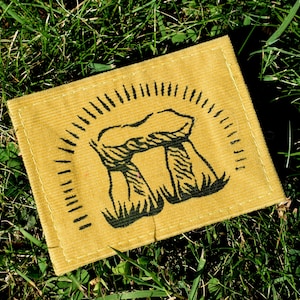 Peut inclure: Un écusson carré jaune moutarde avec une illustration noire d'une structure en pierre, possiblement Stonehenge. Le dessin est entouré d'un motif de rayons de soleil. L'écusson est posé sur de l'herbe verte.