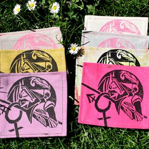 Op de afbeelding: Zes stoffen onderzetters met een zwart-witte grafische print van een vrouwenhoofd met een kroon en een symbool. De onderzetters zijn in verschillende kleuren, waaronder roze, geel en grijs.