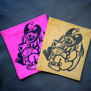 May include: Two square fabric patches. One is bright pink, the other is tan. Each features a black illustration of a seated creature playing a drum. The patches are on a dark gray textured surface.