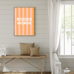 May include: A framed print with pink and orange vertical stripes and the words "BISOUS MORUE" in white. The print is above a wooden bench with patterned cushions. The room has a neutral colour scheme, with white curtains and a wooden floor.