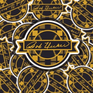 May include: A sticker featuring a black and gold design with the text "Bob Uecker" in a cursive font. The sticker has a circular shape with a banner and baseball-themed accents.