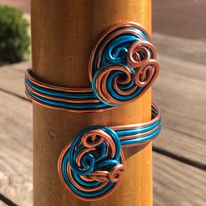 May include: Two copper wire bracelets with blue wire accents. The bracelets are wrapped around a wooden dowel.
