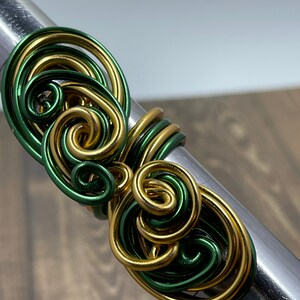 May include: A green and gold wire ring with a swirling design. The ring is made of wire that has been twisted and shaped into a unique pattern.