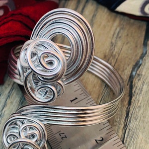 May include: Silver wire ring with a swirling design. The ring is measured against a ruler, showing it is about 2 inches in diameter.