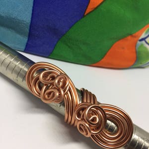 May include: A copper wire ring with a swirling, organic design. The ring is shown on a ruler, measuring approximately 1.5 inches in diameter.