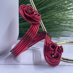 May include: A red wire ornament shaped like a stylized flower with a swirl design. The ornament is made of multiple strands of wire twisted together.