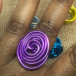 K&ouml;nnte beinhalten: Ein violetter Drahtring mit einem Spiraldesign. Der Ring besteht aus mehreren Drahtwicklungen, die miteinander verdreht sind, um ein einzigartiges und auff&auml;lliges Design zu schaffen.