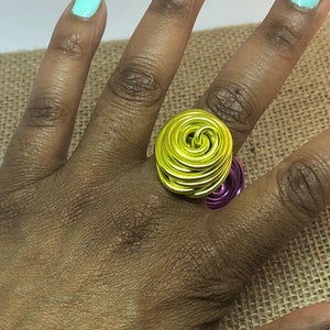 K&ouml;nnte beinhalten: Ein gelber und violetter Drahtring mit einem Spiraldesign. Der Ring wird an einem Finger getragen.