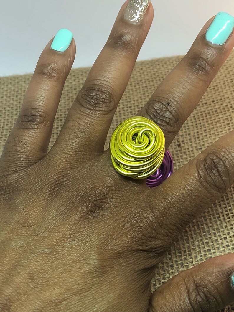 K&ouml;nnte beinhalten: Ein gelber und violetter Drahtring mit einem Spiraldesign. Der Ring wird an einem Finger getragen.