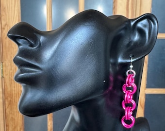 Könnte beinhalten: Ein Hängeohrring mit einer vertikalen Kette aus leuchtend pinkfarbenen Metallringen, die in einem sich wiederholenden Muster verbunden sind. Der Ohrring ist an einem silberfarbenen Haken befestigt und hängt am Ohr einer schwarzen, abstrakten Büste.