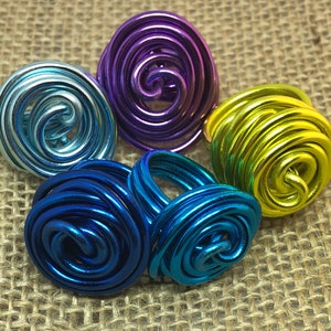 K&ouml;nnte beinhalten: F&uuml;nf farbige Drahtringe, jeder mit einem Spiraldesign. Die Ringe sind in Blaut&ouml;nen, Gr&uuml;n, Lila und T&uuml;rkis.