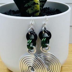 May include: Silver spiral earrings with black and green accents. The earrings feature a spiral design at the bottom, connected by black and green rings to a silver hook. The earrings are displayed in front of a white pot with a green and yellow plant.