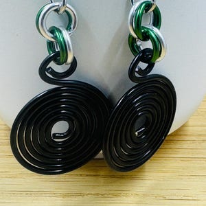 May include: A pair of handmade earrings featuring a spiral design. The earrings have silver-colored hooks and chain links, with green and silver links connecting to a black spiral. The earrings are displayed against a neutral background.
