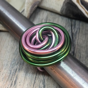 May include: A handmade ring with a floral design. The ring features a woven pattern of pink and green metallic wire. The ring is displayed on a silver metal ring sizer. The background is a light wood surface.