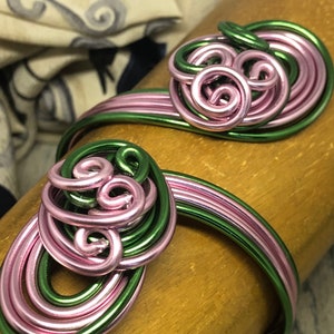 May include: A green and pink wire bracelet with two large, intertwined, spiral designs. The bracelet is made of multiple strands of wire, creating a textured look.