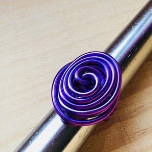 K&ouml;nnte beinhalten: Ein violetter Drahtring mit einem Spiraldesign. Der Ring besteht aus mehreren Drahtwindungen, die miteinander verdreht sind, um ein einzigartiges und auff&auml;lliges Design zu schaffen.